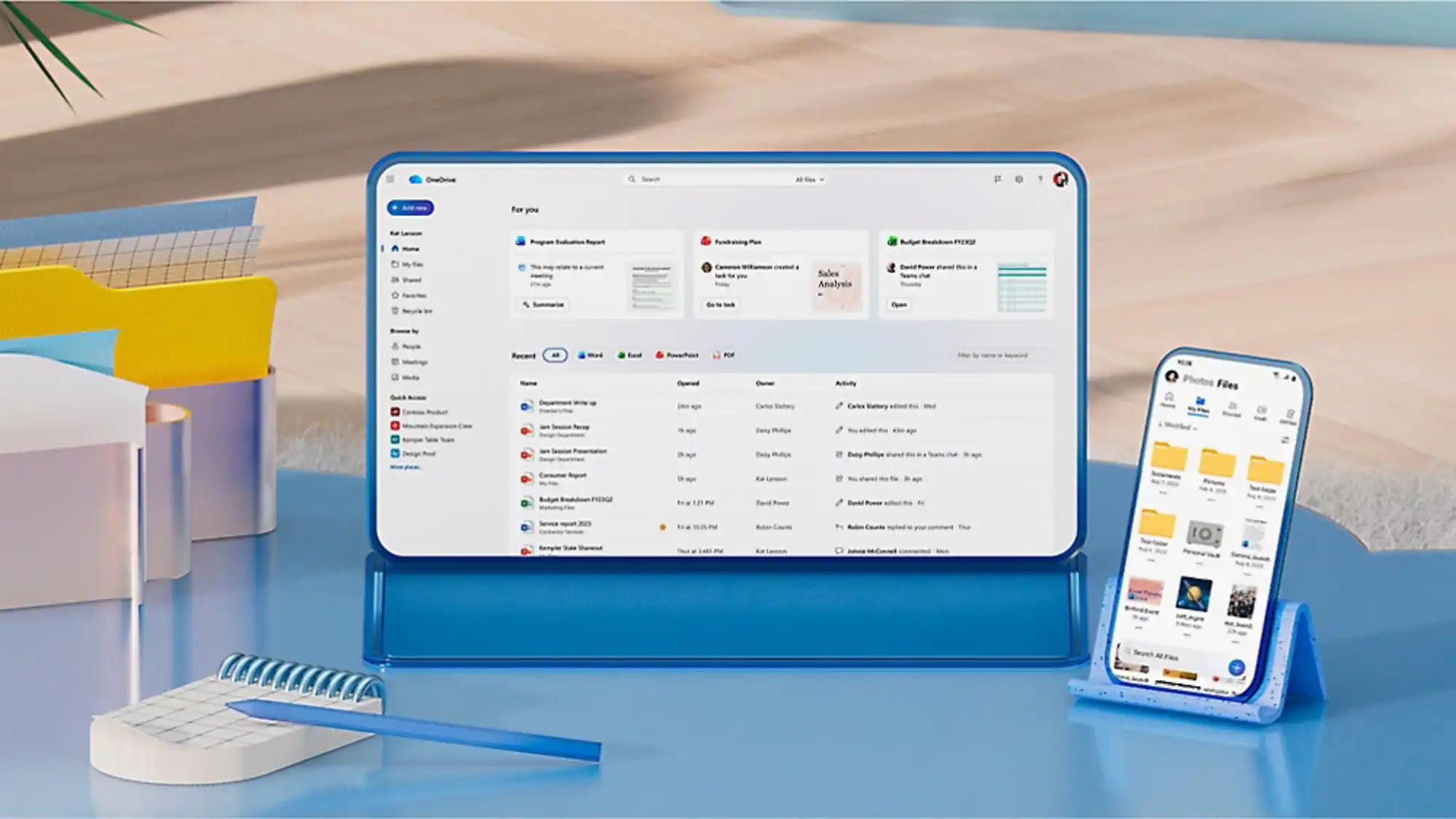 Tablet na niebieskiej podstawce, obok telefon na niebieskiej mniejszej podstawce, na niebieskim stoliku, na beżowym tle podłogi. Na ekranach widać OneDrive, zarówno tabletu jak i telefonu